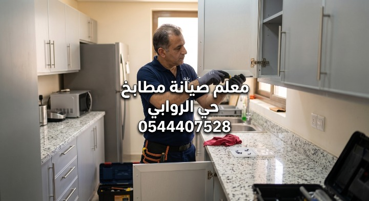 معلم صيانة مطابخ يعمل على إصلاح مفصلة باب دولاب ألمنيوم رمادي. يرتدي الفني قميصا أزرق وقفازات ويستخدم مفك براغي. مكتوب على الصورة: معلم صيانة مطابخ حي الروابي 0544407528. يظهر في الخلفية مطبخ حديث بأسطح رخامية وصندوق أدوات مفتوح.